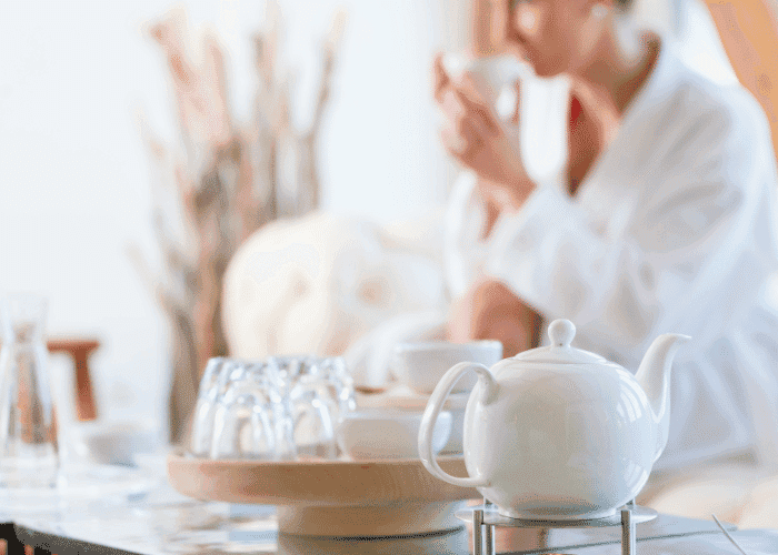 White teapot on table with blurred person in robe drinking tea in background.